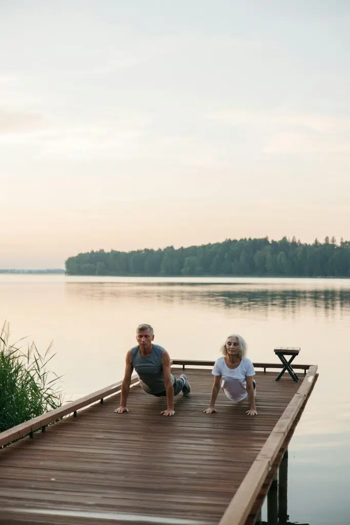 Deux personnes âgées pratiquent le yoga sur un ponton en bois au bord de l’eau