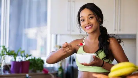 Jeune femme souriante en tenue de sport mangeant un bol de fruits dans une cuisine, illustrant les bienfaits d’un mode de vie actif pour les personnes diabétiques